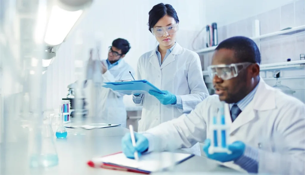 Three scientists in a bright laboratory; a woman in the center holds a blue clipboard while two colleagues conduct experiments nearby.