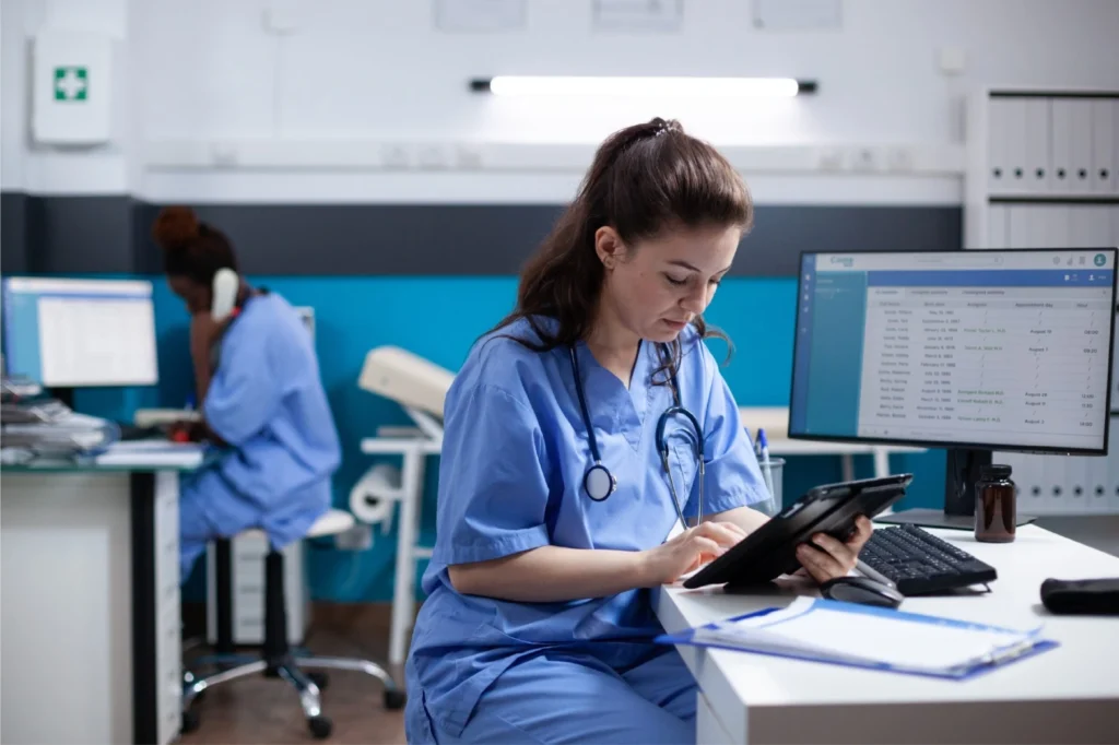 Female nurse in blue scrubs sits at a desk using a tablet, stethoscope around her neck, with a computer monitor nearby.