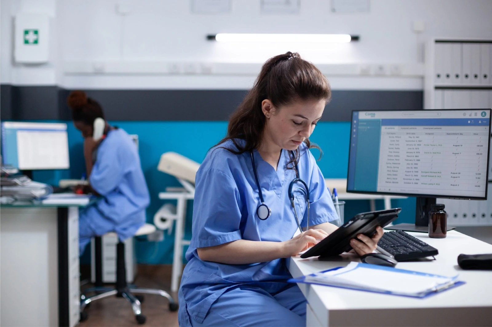 Female nurse in blue scrubs sits at a desk using a tablet, stethoscope around her neck, with a computer monitor nearby.