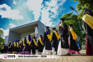 Line of graduates in black gowns with yellow sashes walk outdoors during IIHS Graduation 2026, past a modern building.