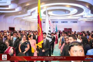 Crowded graduation ceremony with a flag bearer leading a procession down the aisle in a modern hall.