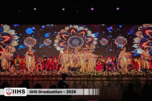Group of dancers performing on a decorated stage for IIHS Graduation 2026, with a vibrant mandala backdrop and seated dignitaries in colorful robes.