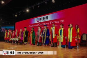 Row of graduates in colorful gowns on stage during IIHS Multiversity Graduation Ceremony 2026, with a red backdrop and banner reading IIHS Multiversity.