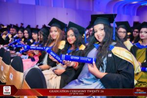 Group of graduates in black gowns and blue stoles, seated and holding blue diploma scrolls at a graduation ceremony.