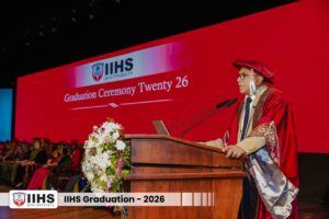 Speaker in red academic robes at a podium during IIHS Multiversity Graduation 2026 ceremony with a flower bouquet nearby on stage.