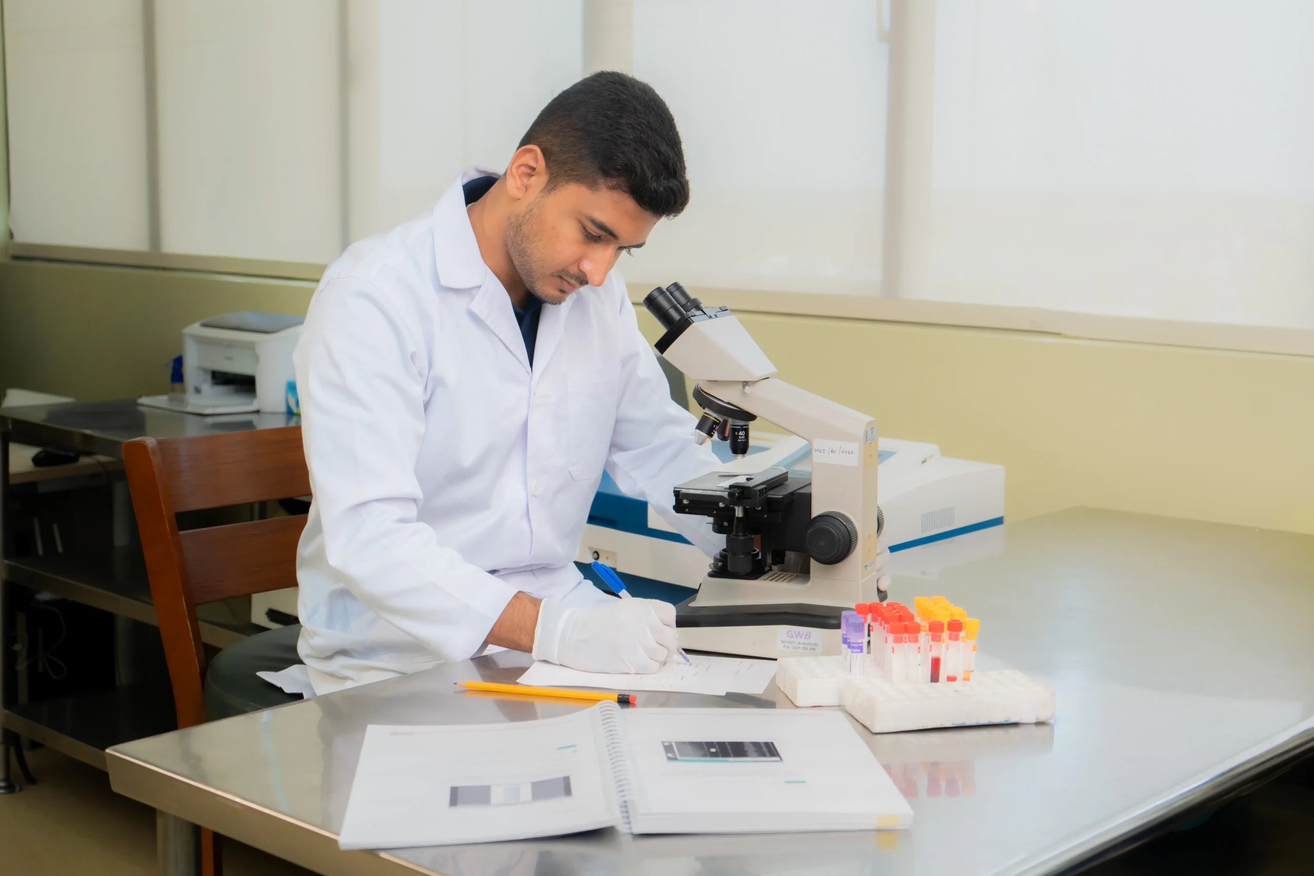 Scientist in a white lab coat examining specimens under a microscope at a lab bench.